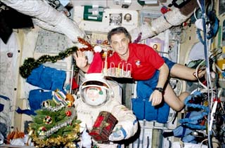 Astronaut David Wolf posing with a santa doll in a Sokol suit, small tree, decked boughs, and floating presents.